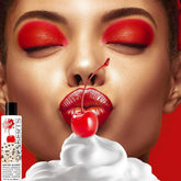 A person wearing vivid red eyeshadow and lipstick gently grips a cherry with their lips above a swirl of whipped cream. To the left, a bottle labeled "Wet Desserts Whipped Cream Flavored Lubricant | 3oz" is paired with chocolate chip cookies, adding a playful twist. The background is an enticing shade of red.